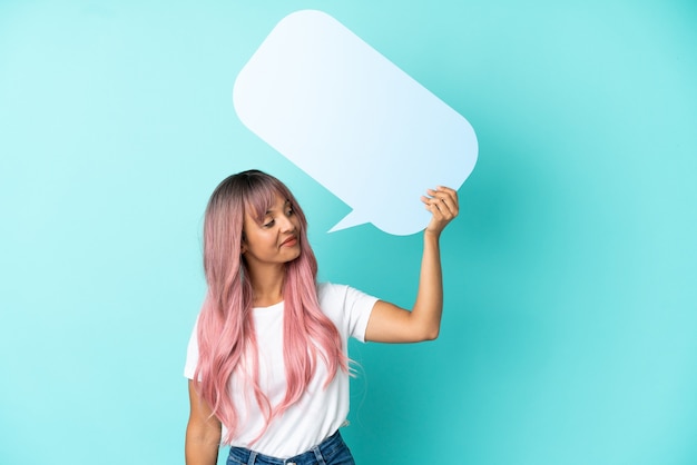 Mulher jovem de raça mista com cabelo rosa isolada em um fundo azul segurando um balão de fala vazio