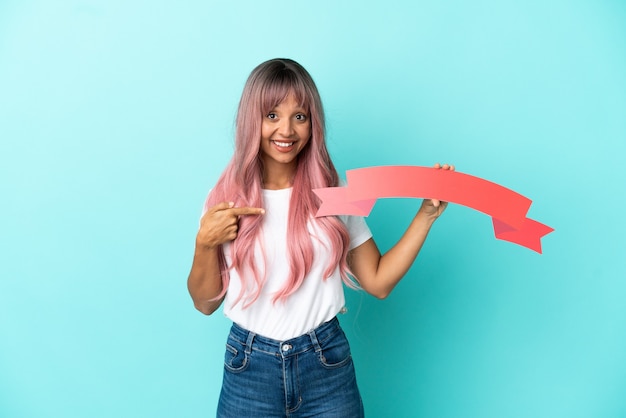 Mulher jovem de raça mista com cabelo rosa isolada em um fundo azul segurando um cartaz vazio com expressão de surpresa