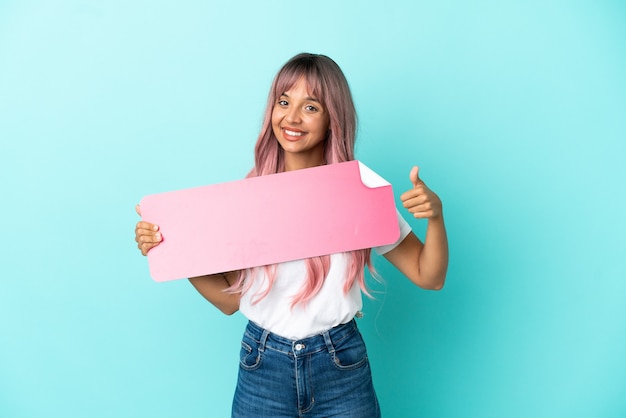 Mulher jovem de raça mista com cabelo rosa isolada em um fundo azul segurando um cartaz vazio com o polegar para cima