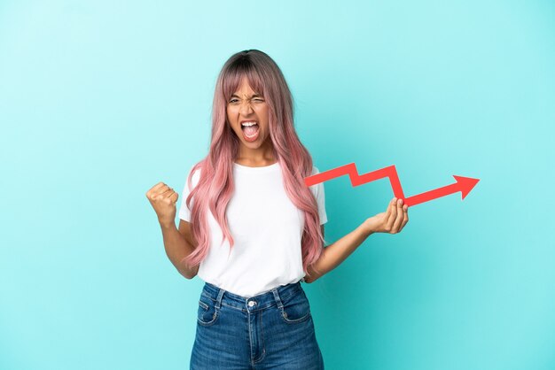 Foto mulher jovem de raça mista com cabelo rosa isolada em um fundo azul, segurando uma flecha ascendente e comemorando uma vitória