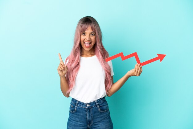 Mulher jovem de raça mista com cabelo rosa isolada em um fundo azul segurando uma seta ascendente e apontando para cima