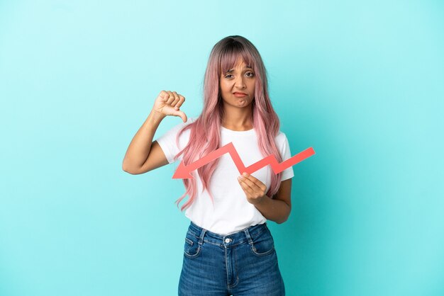 Mulher jovem de raça mista com cabelo rosa isolada em um fundo azul segurando uma seta para baixo e fazendo sinal ruim