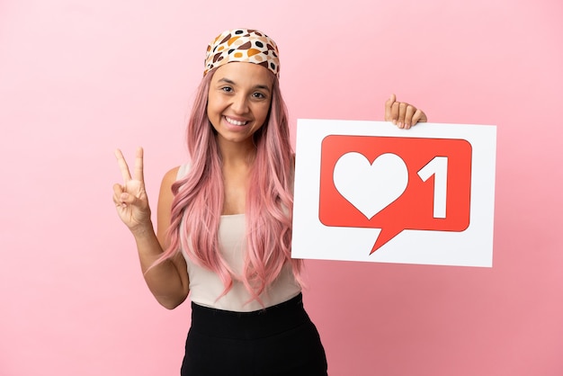 Mulher jovem de raça mista com cabelo rosa isolada em um fundo rosa segurando um cartaz com o ícone de Curtir e comemorando uma vitória