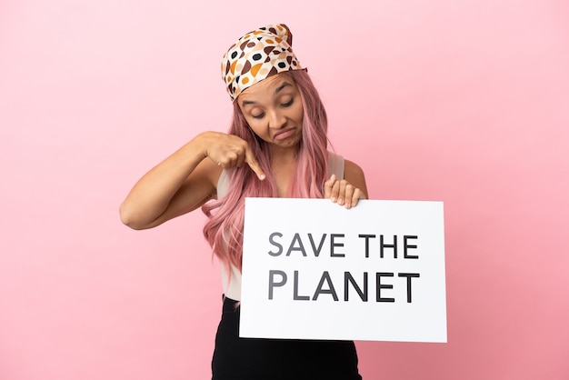 Mulher jovem de raça mista com cabelo rosa isolada em um fundo rosa segurando um cartaz com o texto Salve o Planeta e apontando-o