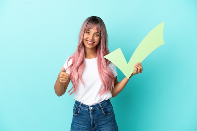 Mulher jovem de raça mista com cabelo rosa isolado em um fundo azul segurando um ícone de seleção com o polegar para cima