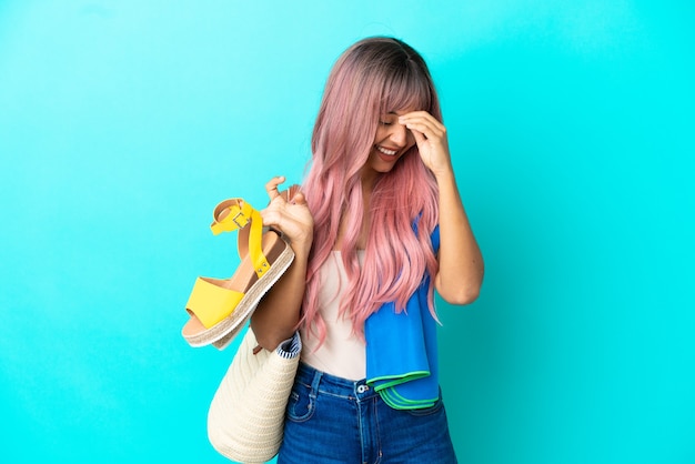 Mulher jovem de raça mista com cabelo rosa segurando sandálias de verão isoladas em um fundo azul rindo