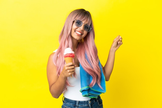 Mulher jovem de raça mista com cabelo rosa segurando sorvete isolado em um fundo amarelo apontando para trás