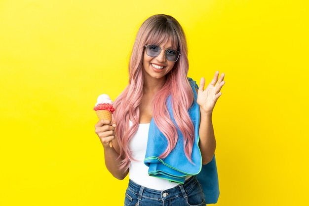 Mulher jovem de raça mista com cabelo rosa segurando um sorvete isolado em um fundo amarelo e saudando com a mão com expressão feliz