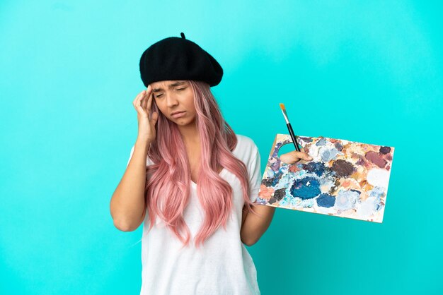 Foto mulher jovem de raça mista com cabelo rosa segurando uma paleta isolada em um fundo azul com dor de cabeça