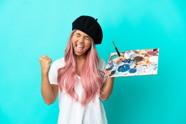 Mulher jovem de raça mista com cabelo rosa segurando uma paleta isolada em um fundo azul comemorando uma vitória