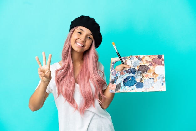 Foto mulher jovem de raça mista com cabelo rosa segurando uma paleta isolada em um fundo azul feliz e contando três com os dedos