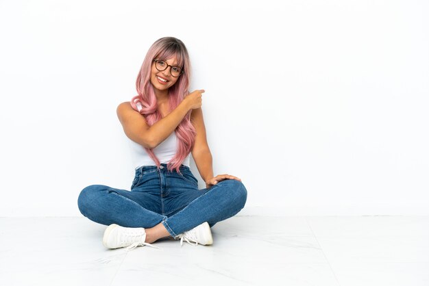 Mulher jovem de raça mista com cabelo rosa sentada no chão isolada no fundo branco apontando para trás