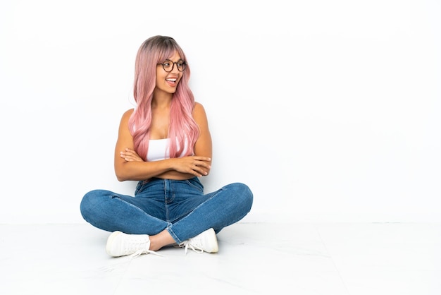 Mulher jovem de raça mista com cabelo rosa sentada no chão, isolada no fundo branco feliz e sorridente