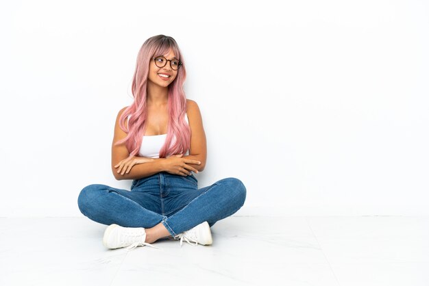 Foto mulher jovem de raça mista com cabelo rosa sentada no chão, isolada no fundo branco, olhando para cima enquanto sorri