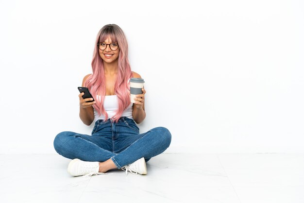 Mulher jovem de raça mista com cabelo rosa sentada no chão, isolada no fundo branco, segurando um café para levar e um celular