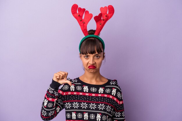 Mulher jovem de raça mista com chapéu de rena, comemorando o Natal, isolado no fundo roxo, mostrando um gesto de antipatia, polegares para baixo. Conceito de desacordo.