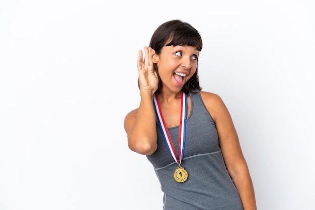 Mulher jovem de raça mista com medalhas isoladas no fundo branco, ouvindo algo, colocando a mão na orelha