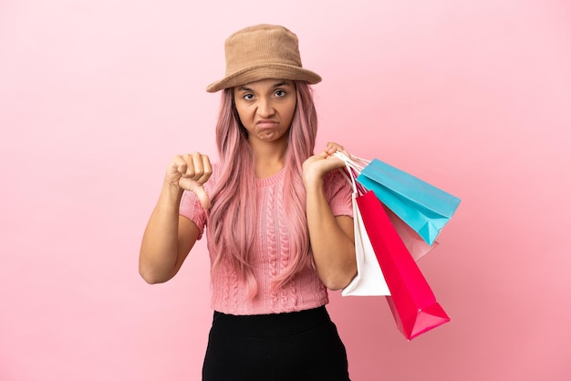 Mulher jovem de raça mista com sacola de compras isolada em fundo rosa, mostrando o polegar para baixo com expressão negativa