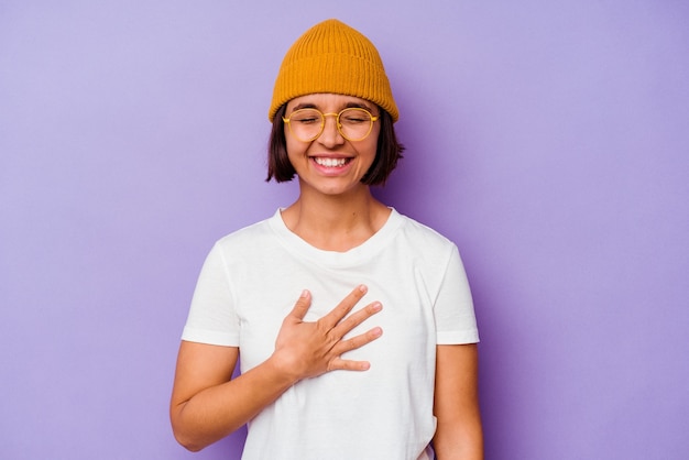 Mulher jovem de raça mista com um boné de lã isolado no fundo roxo ri alto, mantendo a mão no peito.