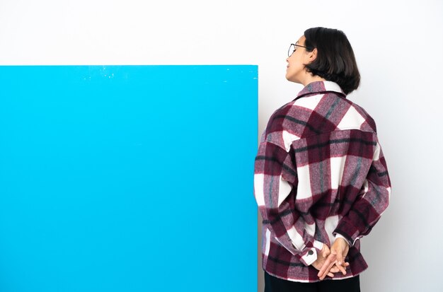 Foto mulher jovem de raça mista com um grande cartaz azul isolado no fundo branco na posição traseira e olhando para trás