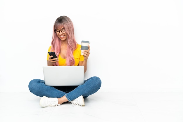 Foto mulher jovem de raça mista com um laptop de cabelo rosa sentada no chão, isolado no fundo branco, segurando um café para levar e um celular