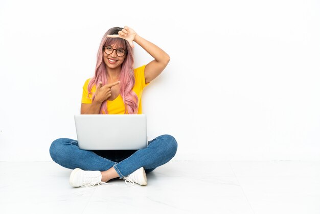 Mulher jovem de raça mista com um laptop de cabelo rosa, sentado no chão, isolado no rosto de foco de fundo branco. Símbolo de enquadramento