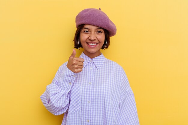 Mulher jovem de raça mista com uma boina isolada em um fundo amarelo, sorrindo e levantando o polegar