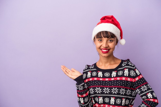 Mulher jovem de raça mista comemorando o natal isolado no fundo roxo, mostrando um espaço de cópia na palma da mão e segurando a outra mão na cintura.