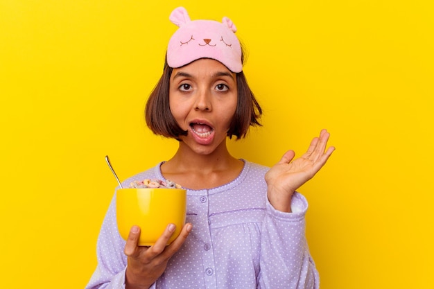 Mulher jovem de raça mista comendo cereais vestindo um pijama isolado na parede amarela surpresa e chocada.