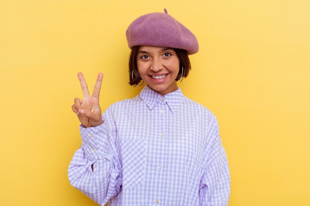 Mulher jovem de raça mista de boina isolada em fundo amarelo, mostrando sinal de vitória e sorrindo amplamente.