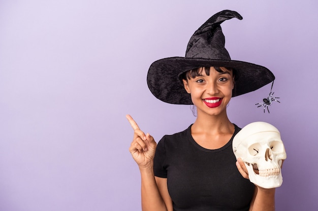 Mulher jovem de raça mista disfarçada de bruxa, segurando uma caveira isolada no fundo roxo, sorrindo e apontando para o lado, mostrando algo no espaço em branco.