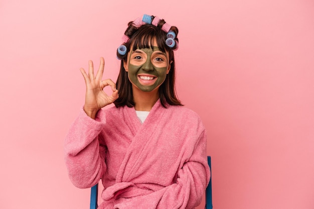 Mulher jovem de raça mista ficando bonita em uma casa isolada em fundo rosa alegre e confiante, mostrando o gesto okey