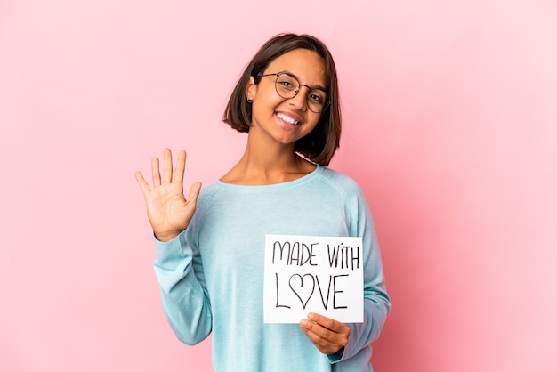 Mulher jovem de raça mista hispânica segurando um cartaz de papel feito com amor sorrindo alegre mostrando o número cinco com os dedos.