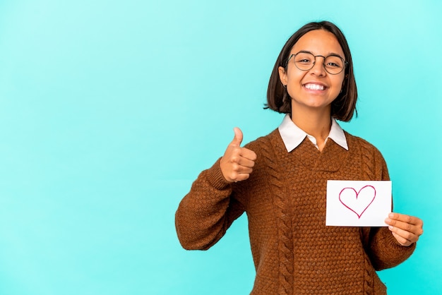 Mulher jovem de raça mista hispânica segurando um papel de coração sorrindo e levantando o polegar