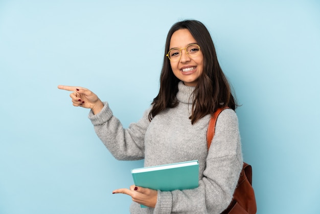 Mulher jovem de raça mista, indo para a escola isolada na parede azul, apontando o dedo para o lado e apresentando um produto