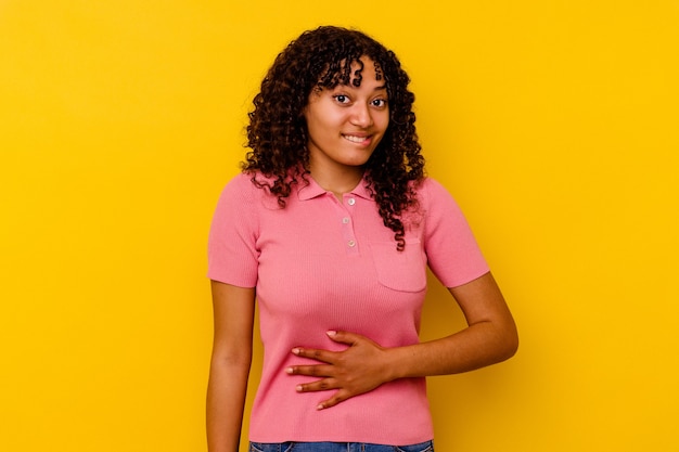 Mulher jovem de raça mista isolada em amarelo toca a barriga, sorri suavemente, conceito de comer e satisfação.