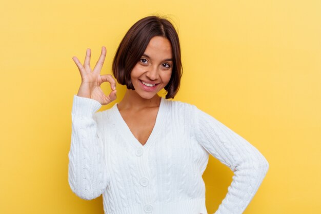 Mulher jovem de raça mista isolada em fundo amarelo alegre e confiante, mostrando um gesto ok.
