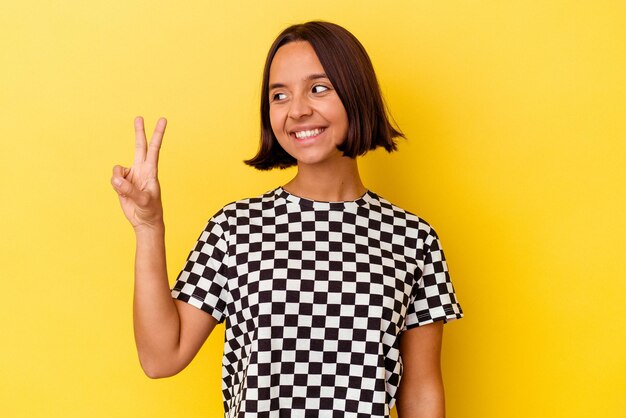 Mulher jovem de raça mista isolada em fundo amarelo alegre e despreocupada, mostrando um símbolo de paz com os dedos.