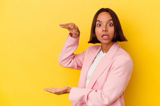 Mulher jovem de raça mista isolada em fundo amarelo chocada e espantada segurando um espaço de cópia entre as mãos.