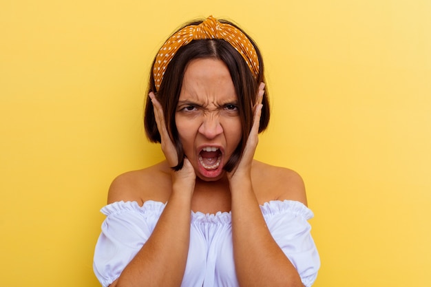 Mulher jovem de raça mista isolada em fundo amarelo, cobrindo as orelhas com as mãos, tentando não ouvir o som muito alto.