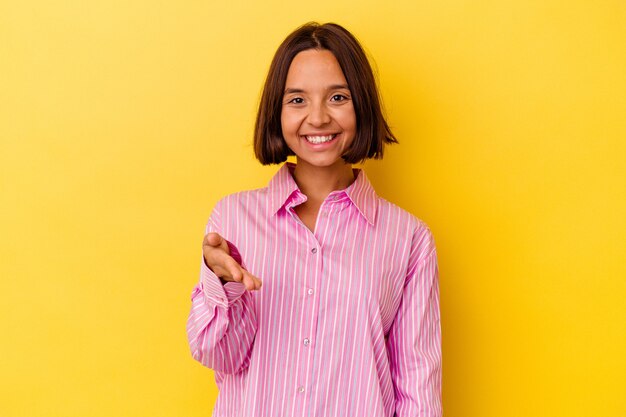 Mulher jovem de raça mista isolada em fundo amarelo, esticando a mão na câmera em gesto de saudação.