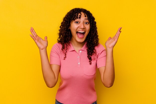 Mulher jovem de raça mista isolada em fundo amarelo, recebendo uma agradável surpresa, animada e levantando as mãos.
