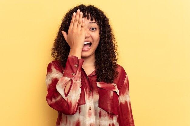 Mulher jovem de raça mista isolada em fundo amarelo, se divertindo, cobrindo metade do rosto com a palma da mão.