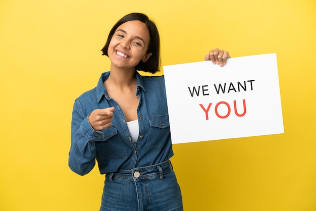 Mulher jovem de raça mista isolada em fundo amarelo segurando a placa We Want You e apontando para a frente