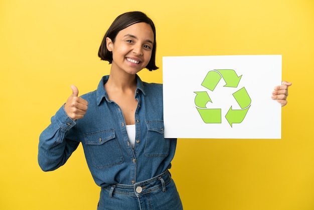 Mulher jovem de raça mista isolada em fundo amarelo segurando um cartaz com o ícone de reciclagem com o polegar para cima