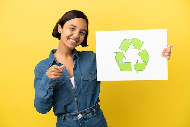 Mulher jovem de raça mista isolada em fundo amarelo segurando um cartaz com o ícone de reciclagem e apontando para a frente