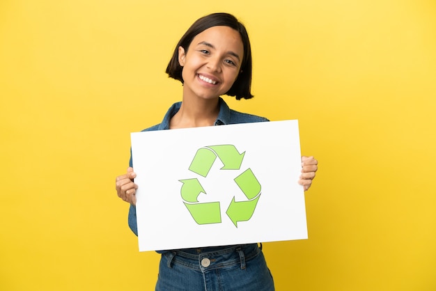 Mulher jovem de raça mista isolada em fundo amarelo segurando um cartaz com o ícone de reciclar e uma expressão feliz