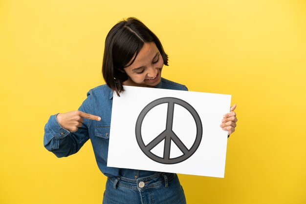 Mulher jovem de raça mista isolada em fundo amarelo segurando um cartaz com o símbolo da paz e apontando-o