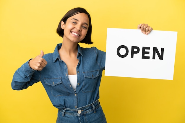 Mulher jovem de raça mista isolada em fundo amarelo segurando um cartaz com o texto ABRIR com o polegar para cima