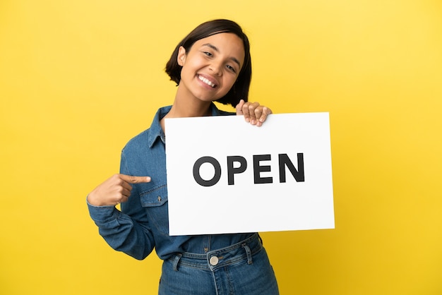 Mulher jovem de raça mista isolada em fundo amarelo segurando um cartaz com o texto ABRIR e apontando-o